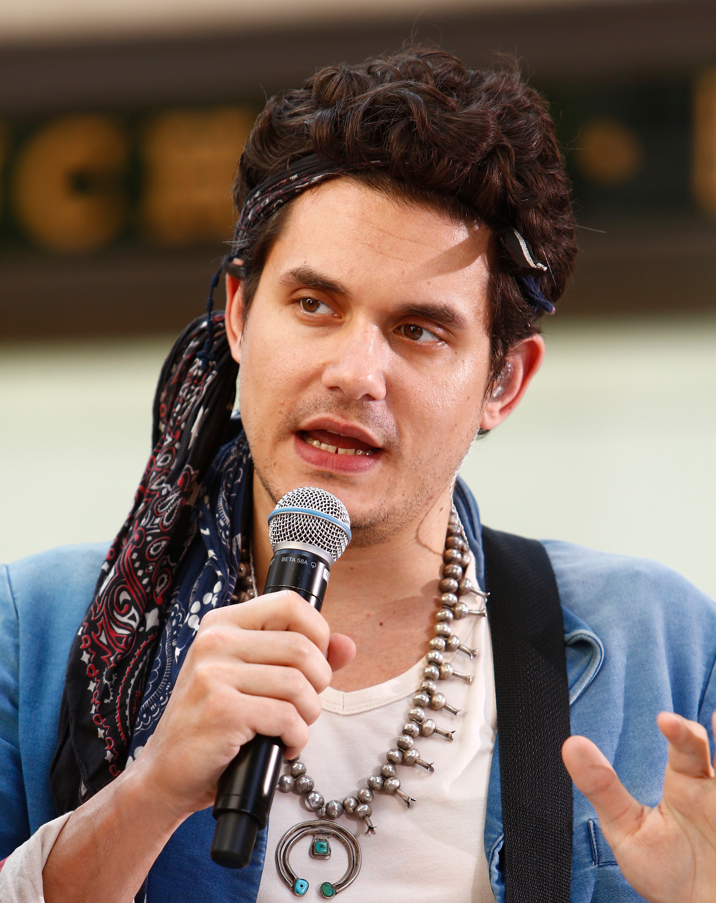 A man with a microphone wearing a bandana and layered necklaces is speaking at an event