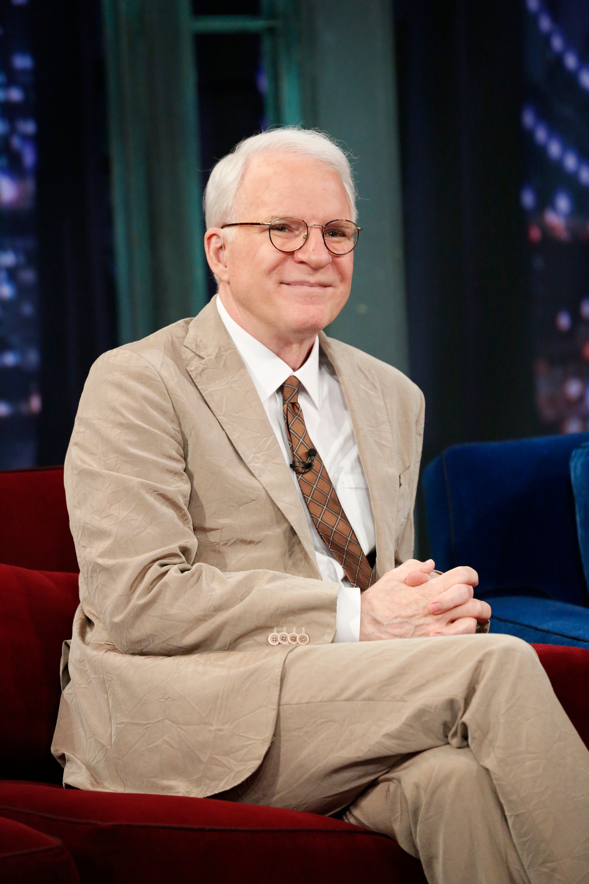 Person in a crumpled suit, seated on a talk show set, smiling with hands clasped