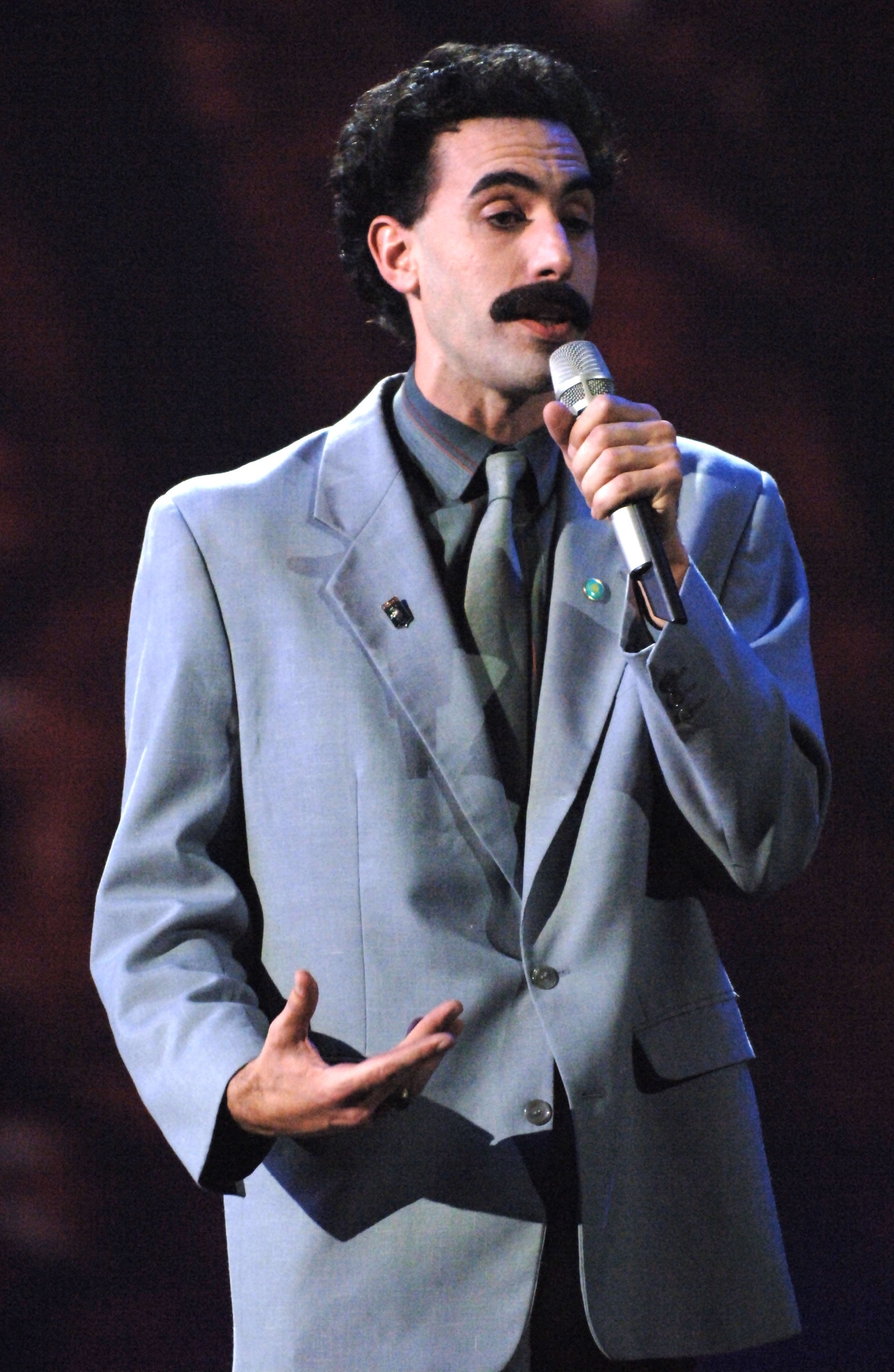 Person in a suit with a mustache holding a microphone, appearing to speak on stage