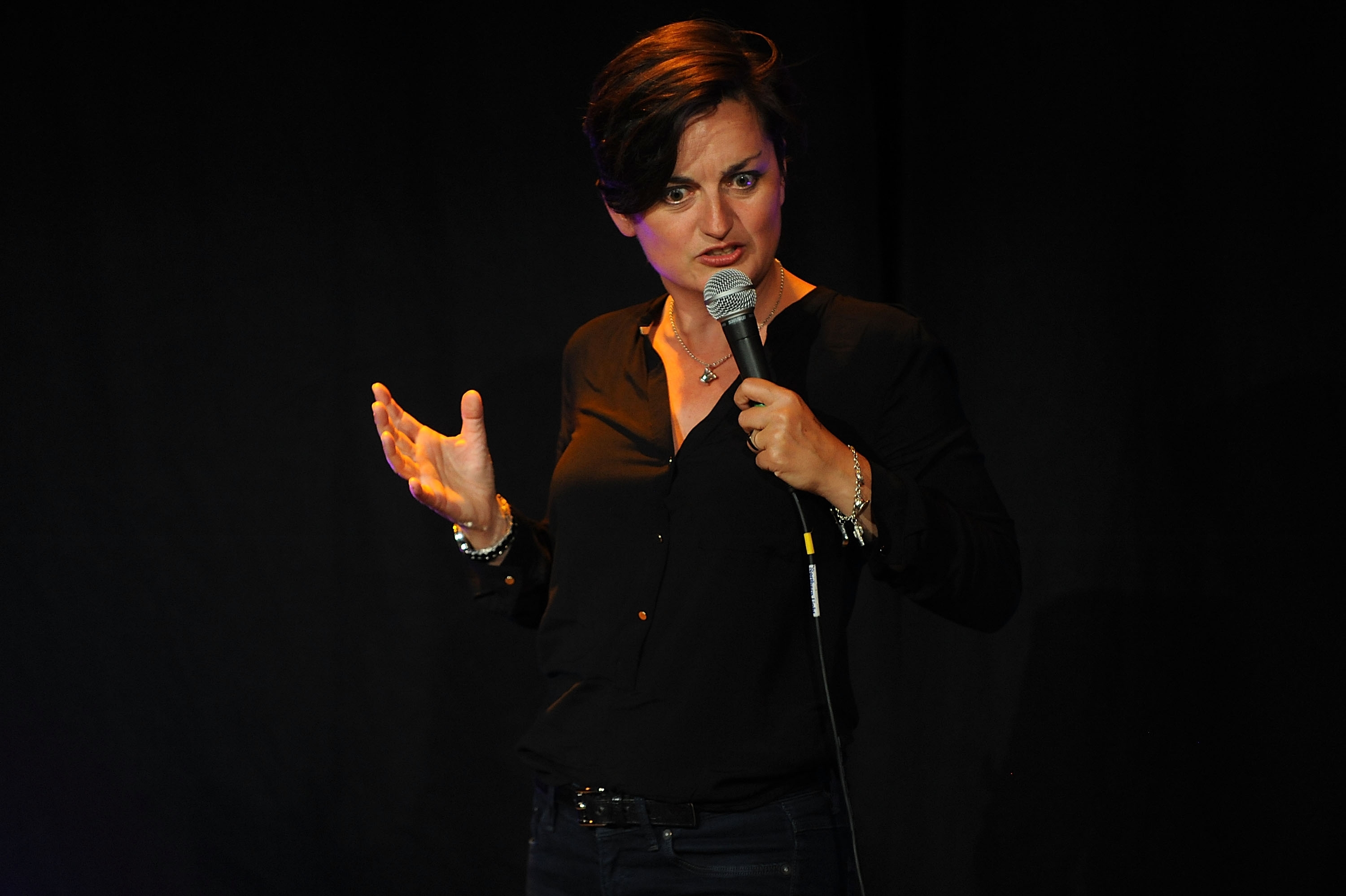 Person on stage in a casual black blouse, holding a microphone, performing stand-up comedy
