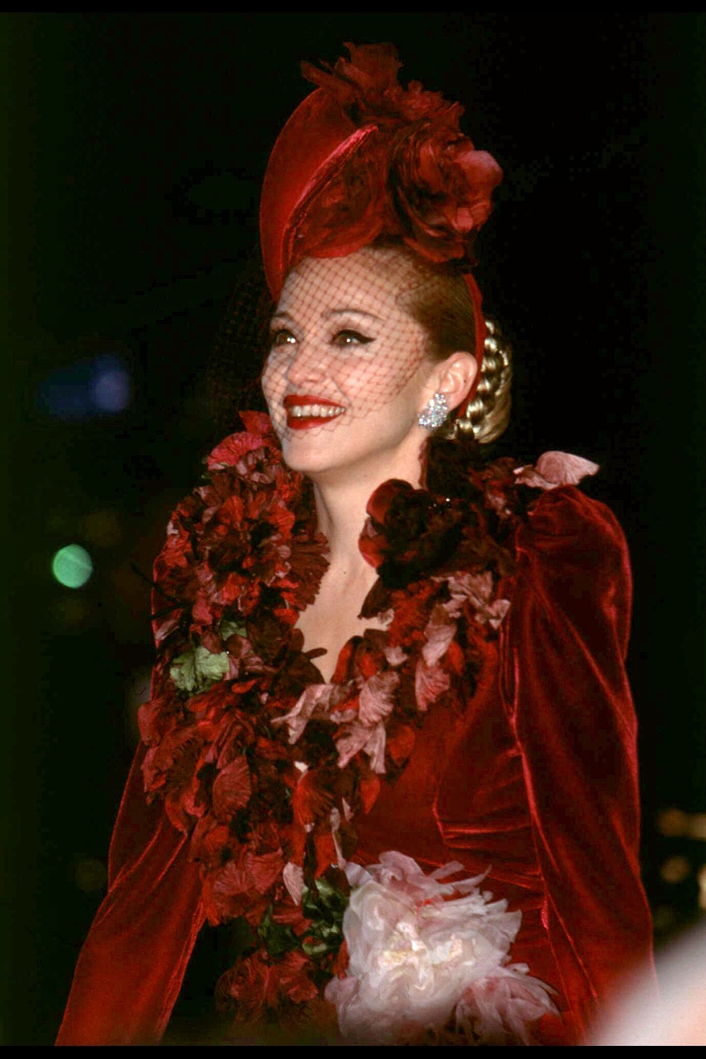 Person in vintage-inspired attire with floral accessories and netted hat on red carpet
