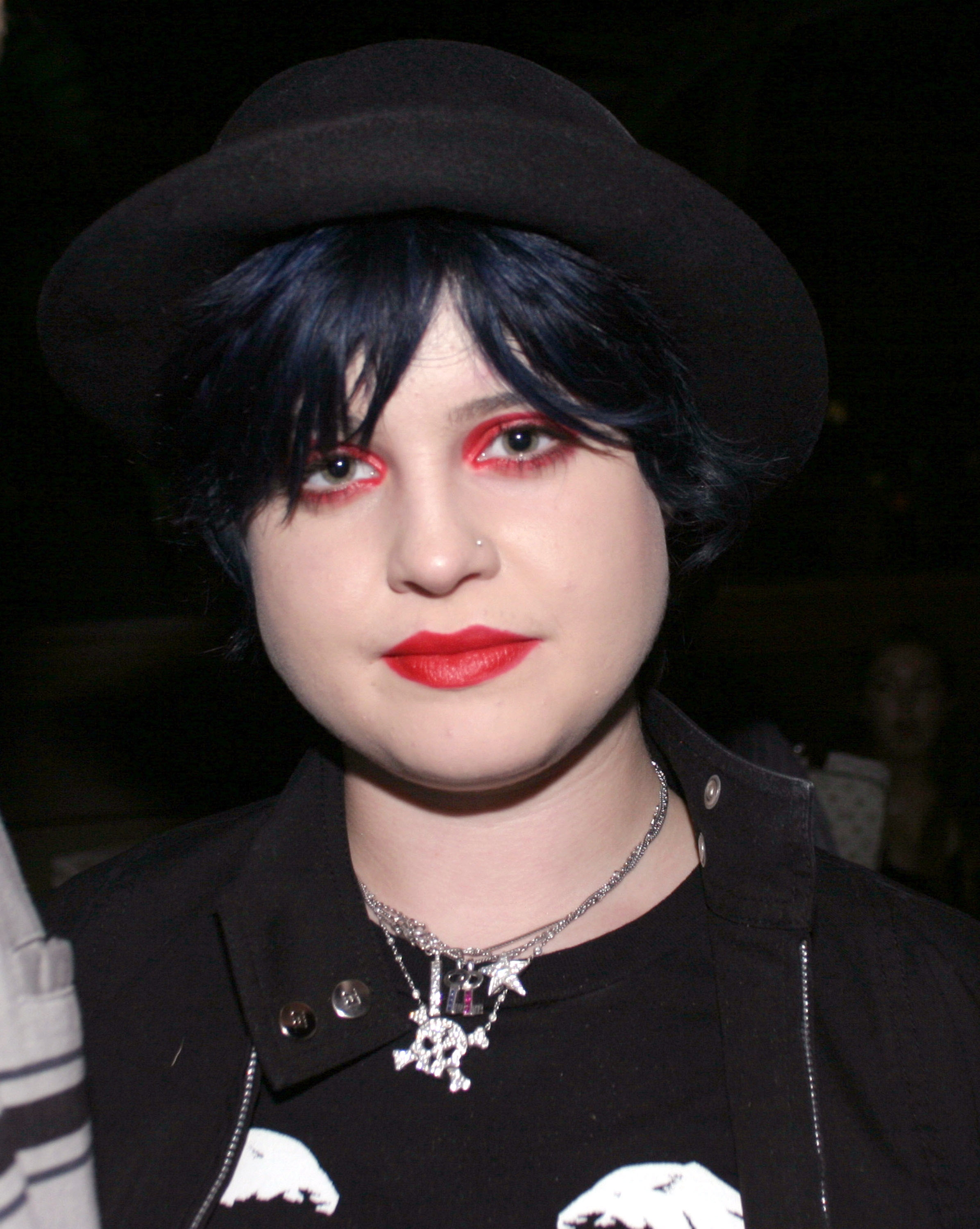 Person in dark hat and jacket, with bold makeup and layered necklaces, attending a celebrity event