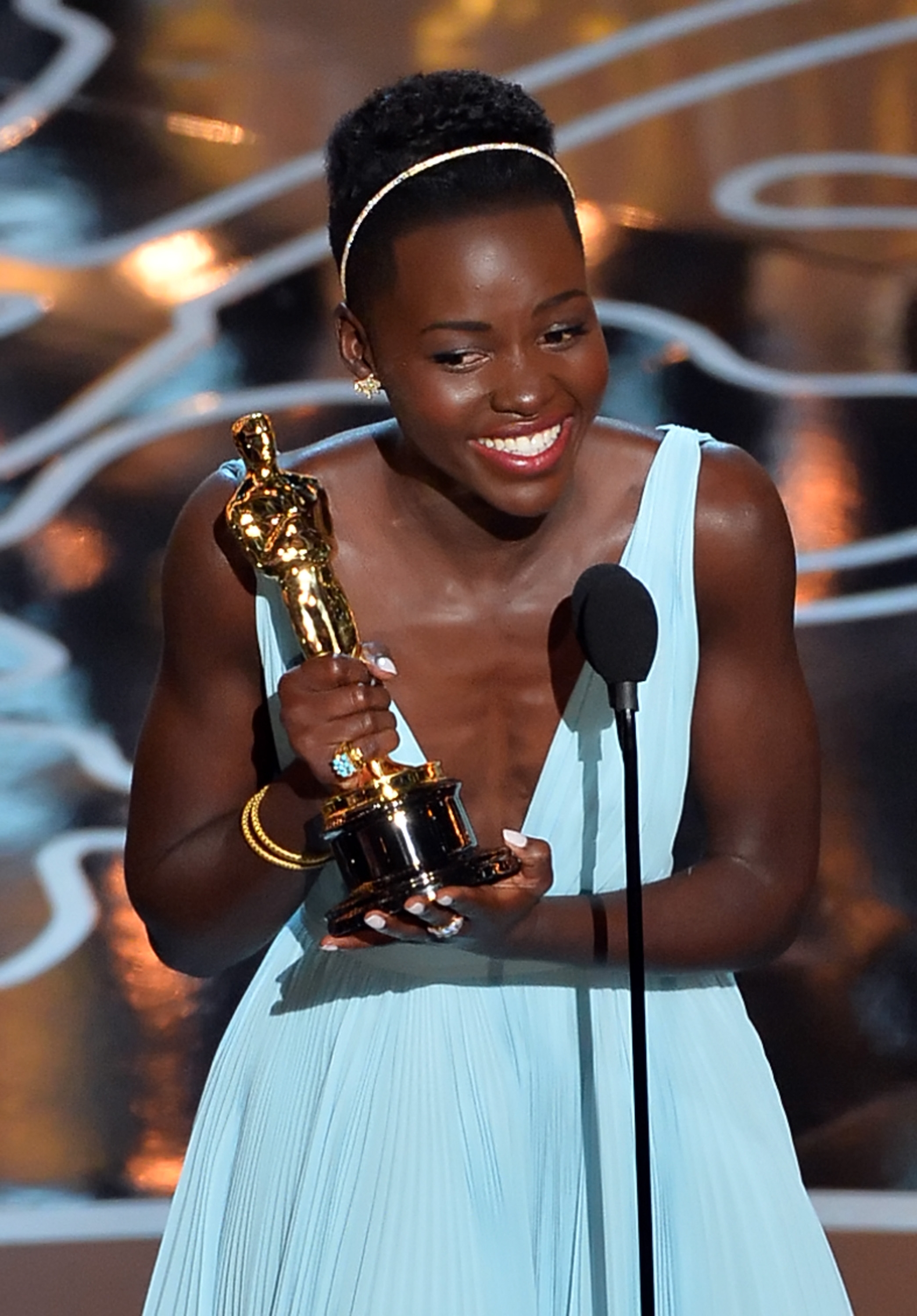 A person in an elegant pleated gown holds an award statue while smiling on stage