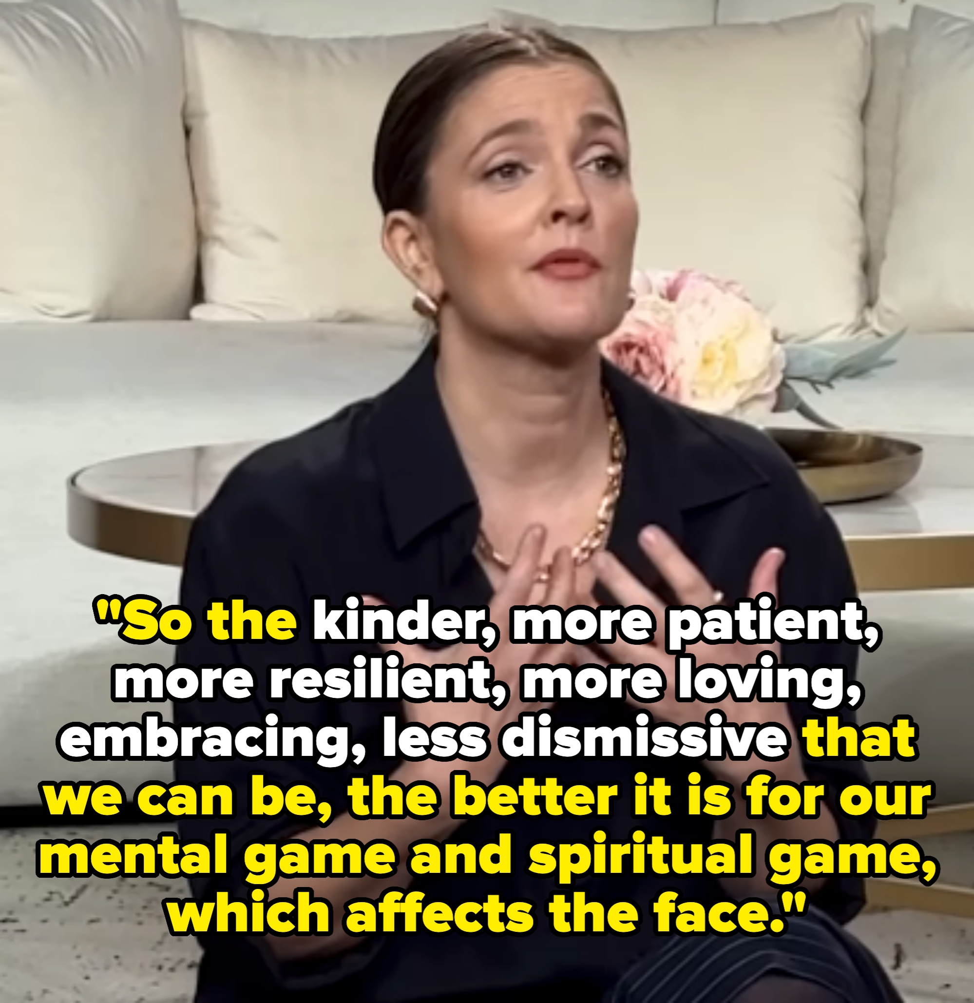 Person seated indoors, gesturing expressively with hands on chest, wearing a black top and necklace against a soft, neutral backdrop