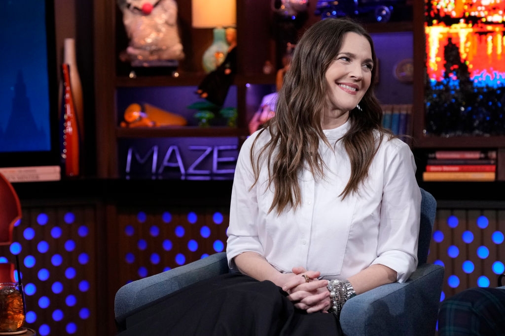Person smiling, seated on a talk show set, wearing a white blouse and black skirt. The background features colorful lights and decor