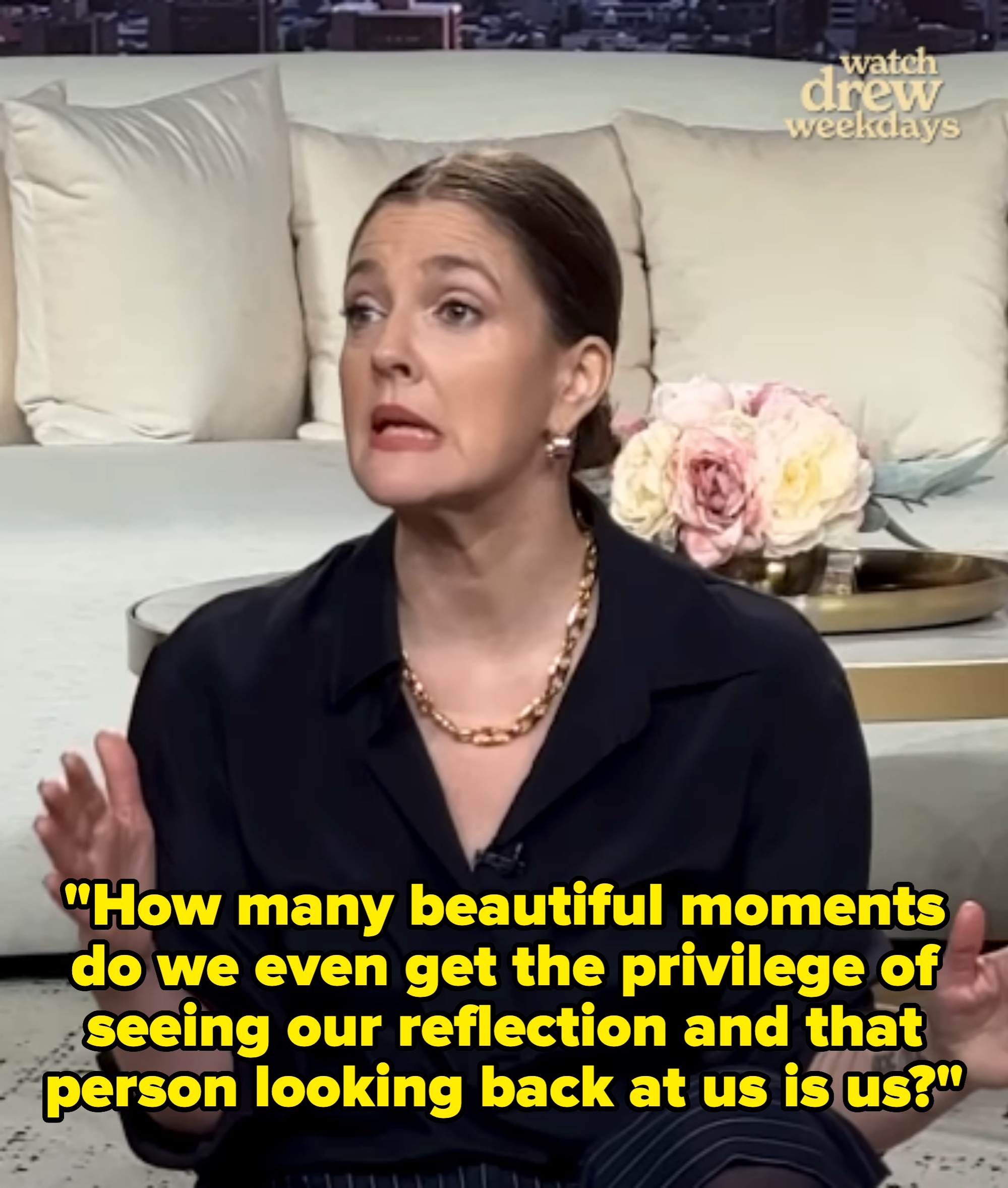 Person speaking on a talk show set, wearing a dark shirt and gold necklace, gesturing with hands