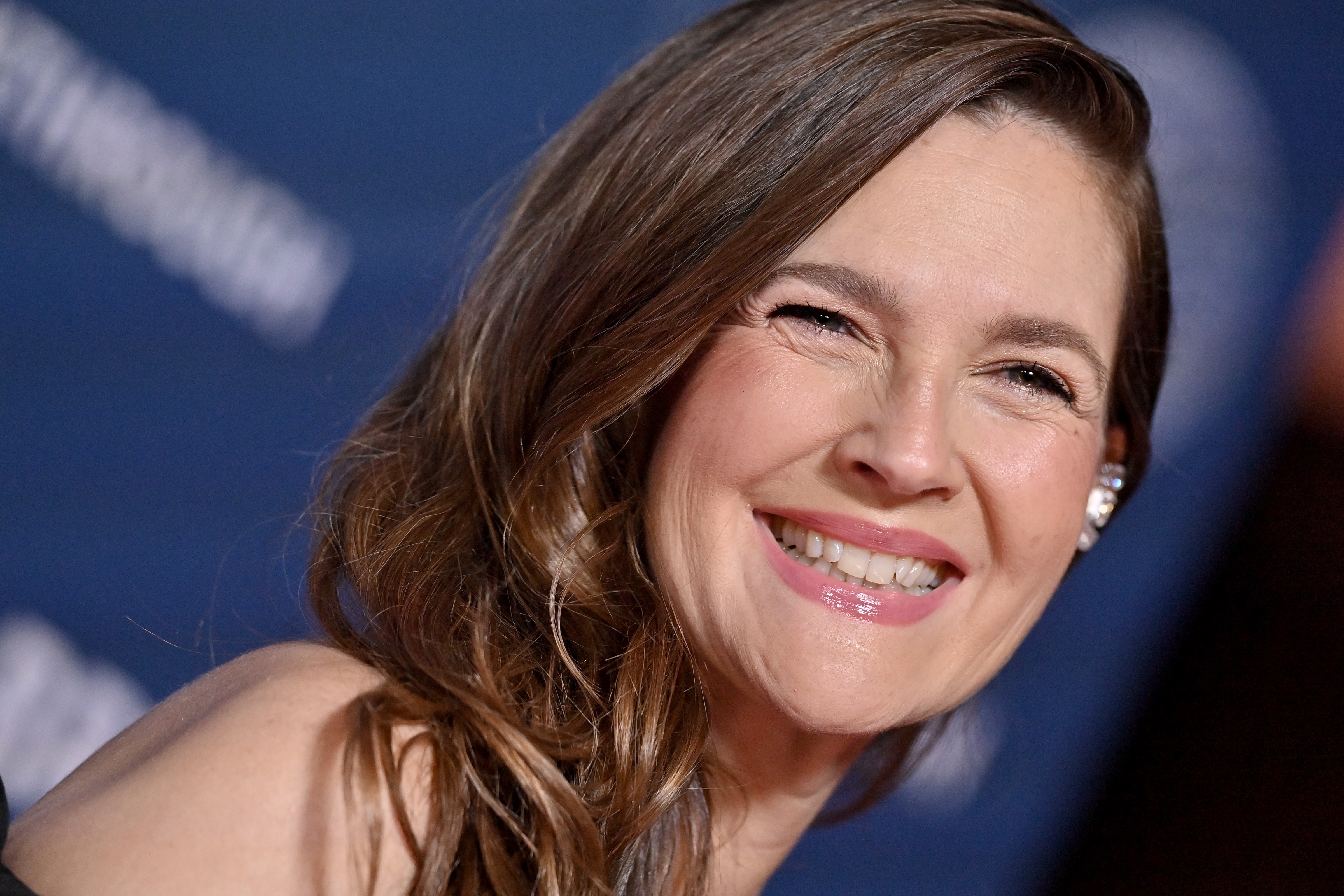 Person smiling at an event. They have long hair and are wearing an off-shoulder outfit with earrings