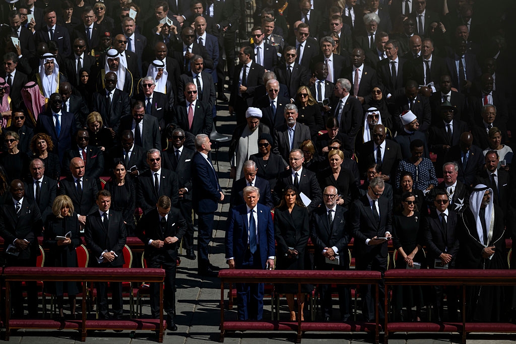 A large group of people in formal black attire attend an outdoor event, seemingly a ceremony or gathering, with some figures standing prominently in the middle
