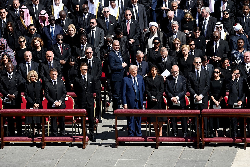 A group of formally dressed individuals stand solemnly at a public event, with some wearing suits and ties