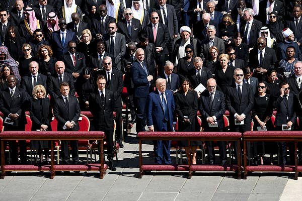 A group of formally dressed individuals stand solemnly at a public event, with some wearing suits and ties