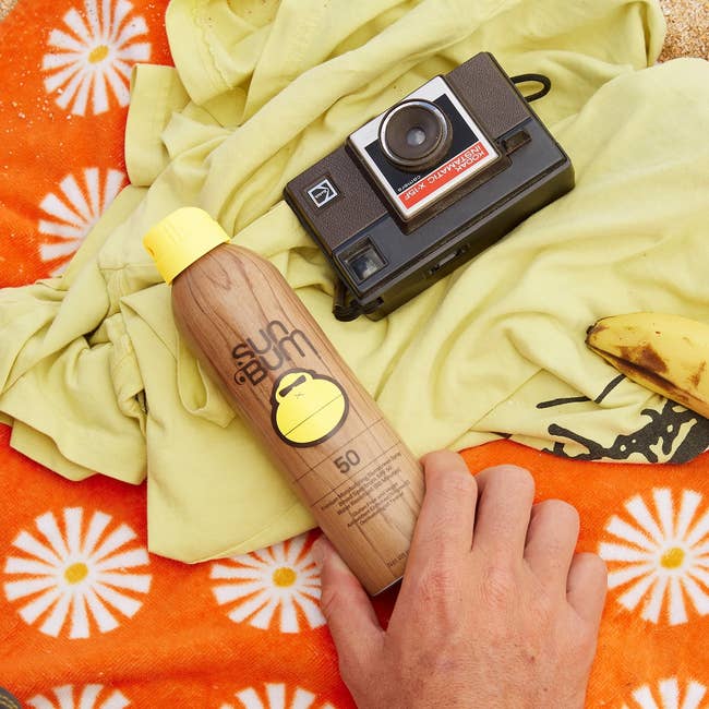 Sunscreen spray, and camera connected a patterned towel astatine the beach. Hand holds the sunscreen