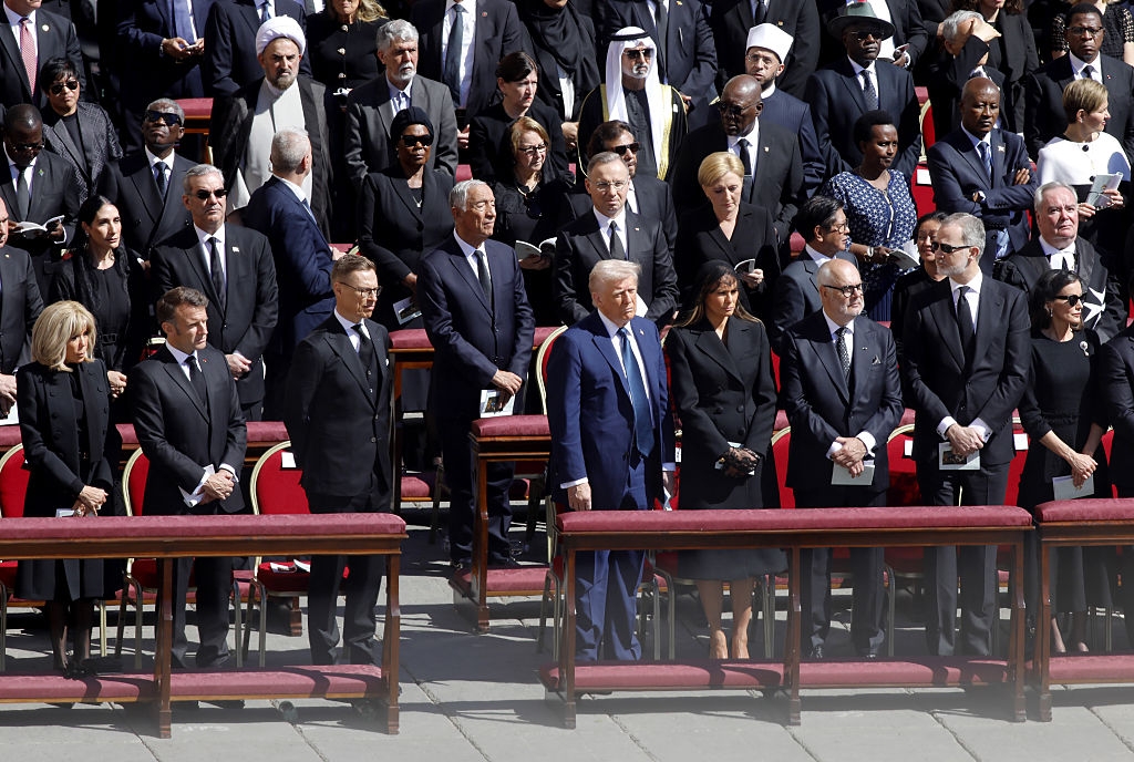 A group of dignitaries stand solemnly in formal attire at an outdoor event, surrounded by an audience