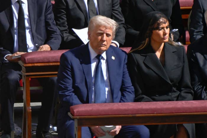 Two individuals seated at an event, dressed in formal attire. The person on the right is wearing a black outfit and headpiece