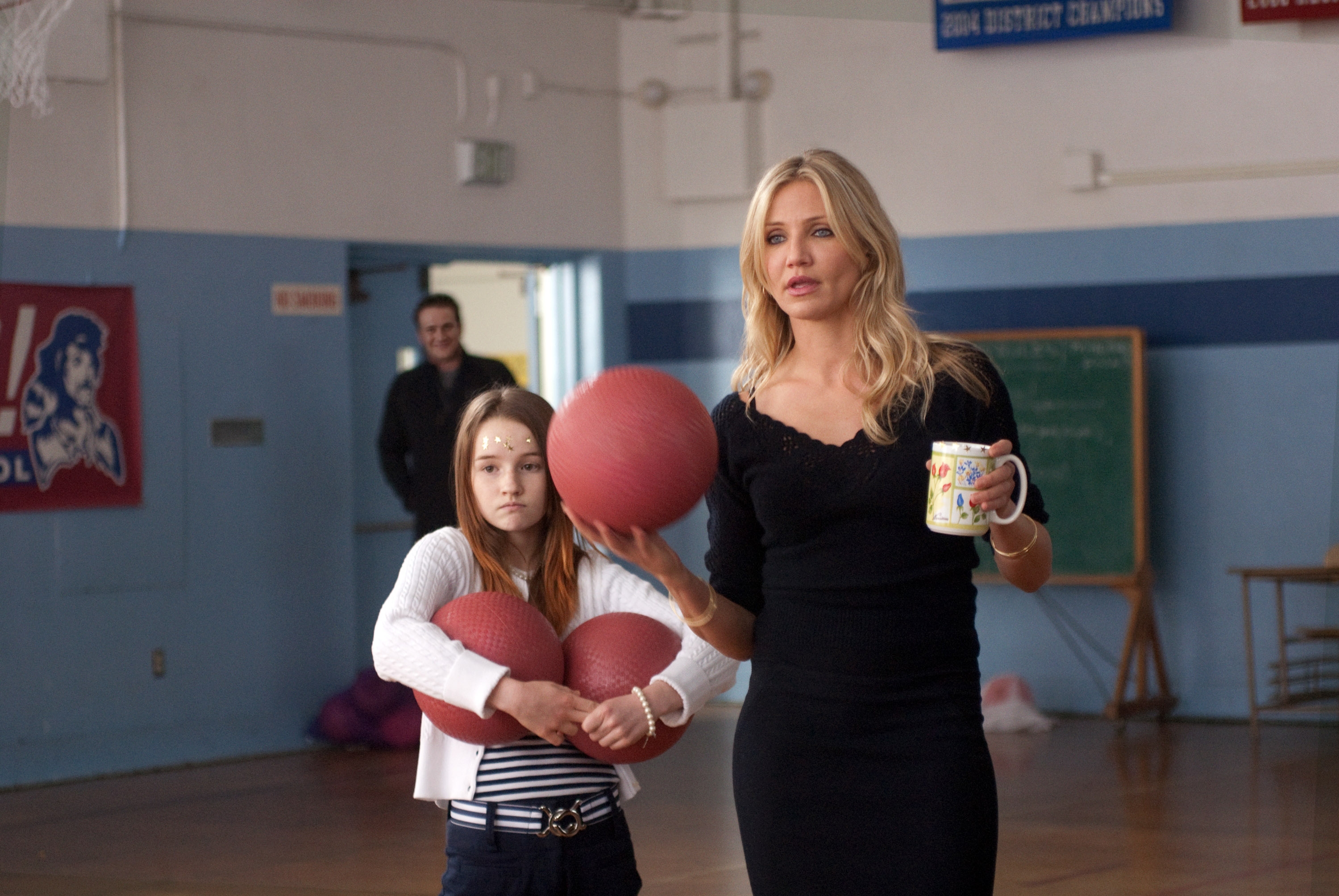 A woman holds a mug and a dodgeball, standing beside a child holding two dodgeballs, in a gym setting