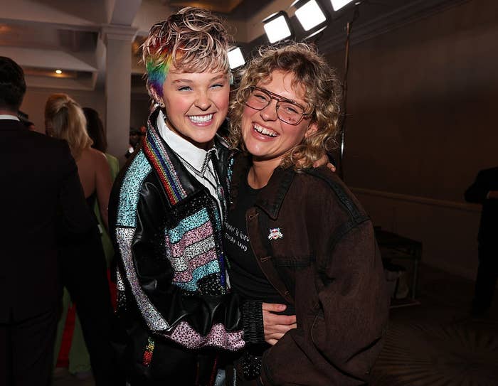 Two people smiling and embracing at an event. One wears a sequined jacket with rainbow detail, the other wears glasses and a casual ensemble