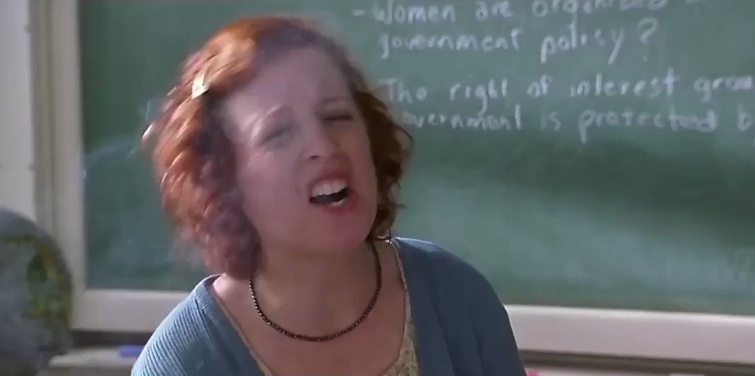 Person in classroom scene speaking passionately in front of a chalkboard with partially visible text