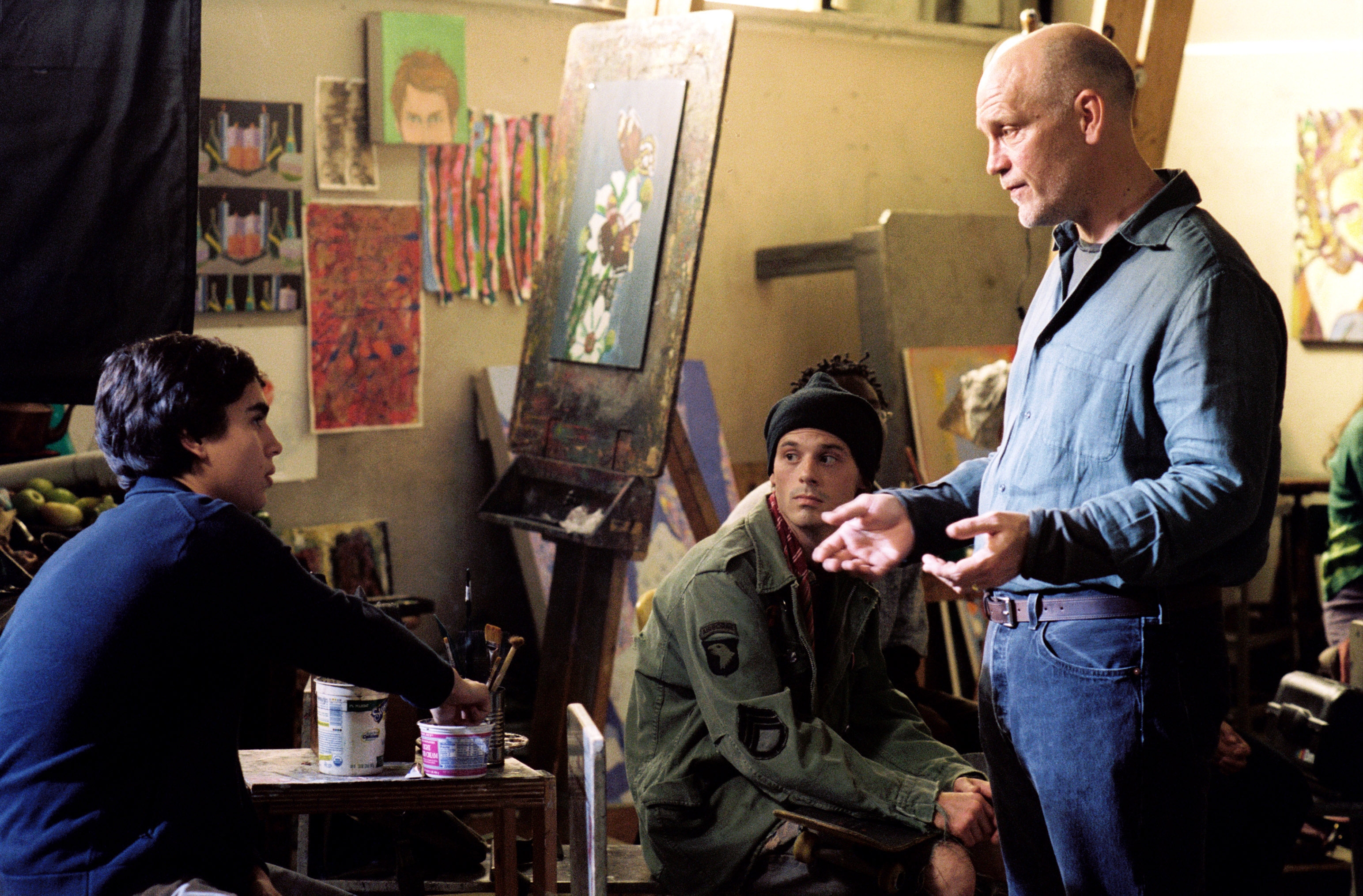 Three men converse in an art studio surrounded by paintings and supplies