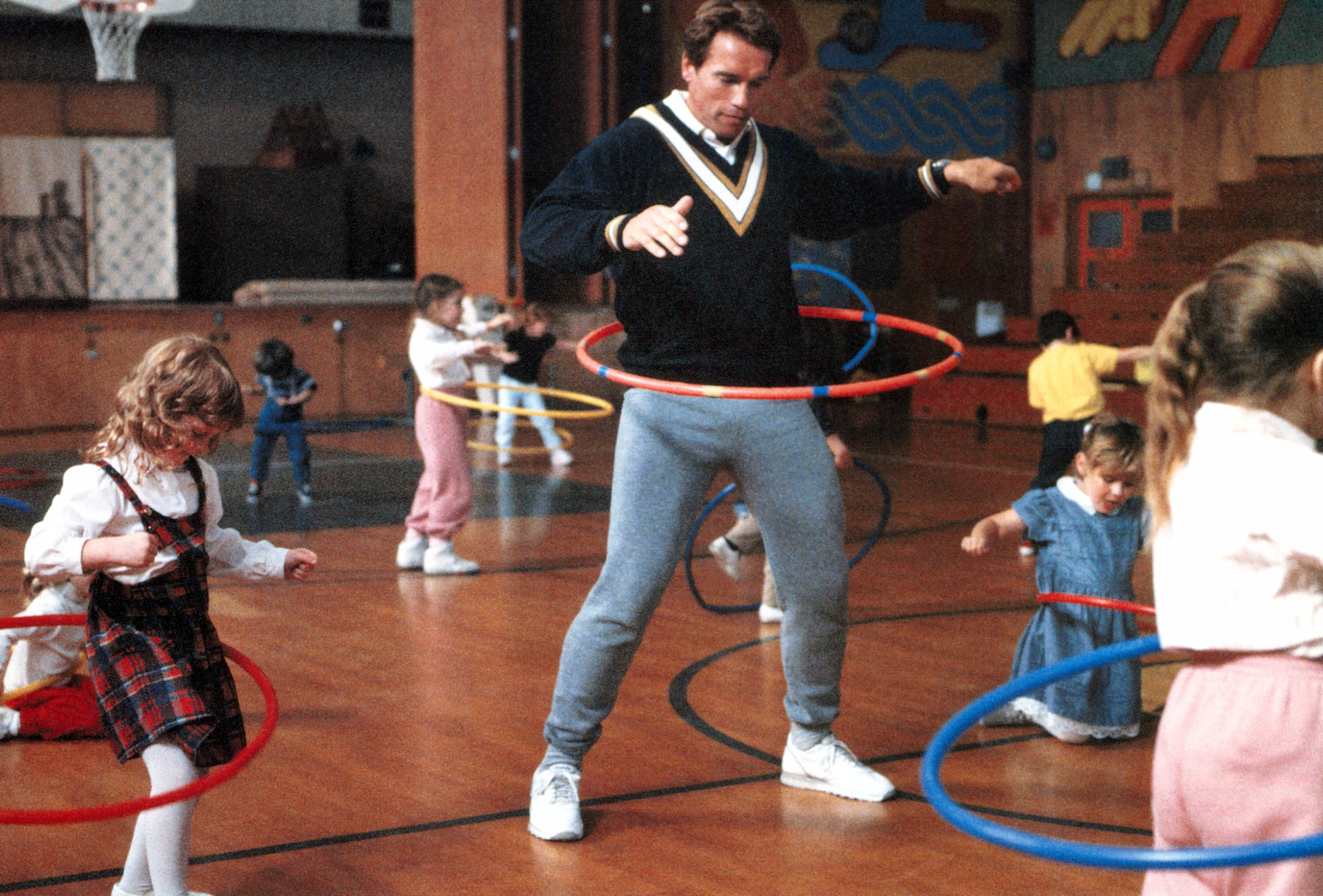An adult and children hula hoop in a gym. The adult wears a sweater and sweatpants, joining kids in playful exercise