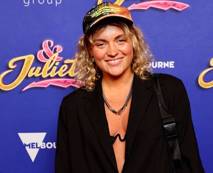 Person in casual jacket, cap, and necklace at a red carpet event, standing in front of an "& Juliet" backdrop.