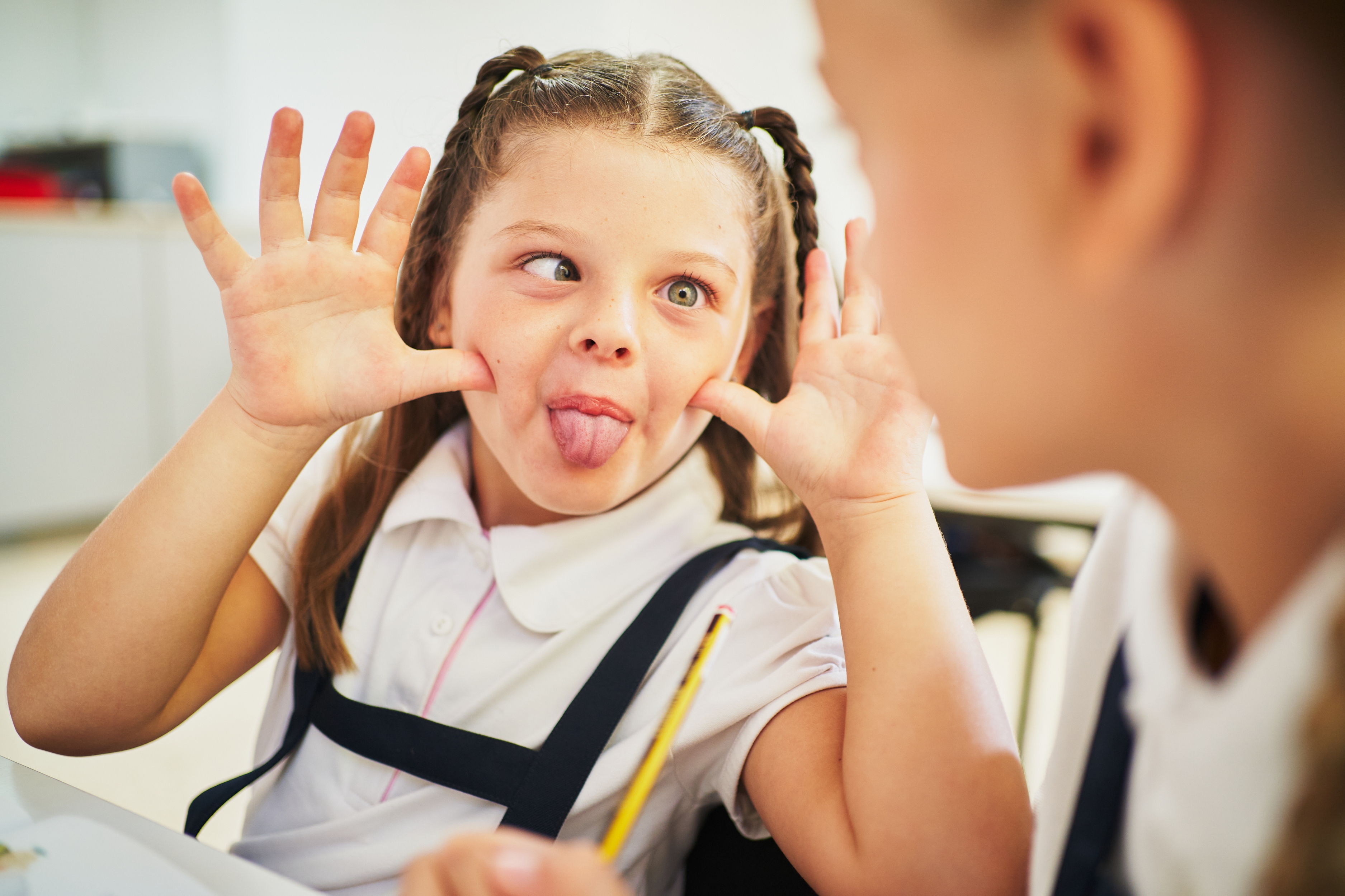 Young girl in school uniform playfully making a silly face with her tongue out, facing another child in the foreground