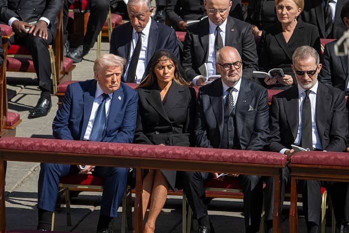 People seated at an outdoor event in formal attire, appearing solemn. They are surrounded by others in similar attire, suggesting a formal gathering