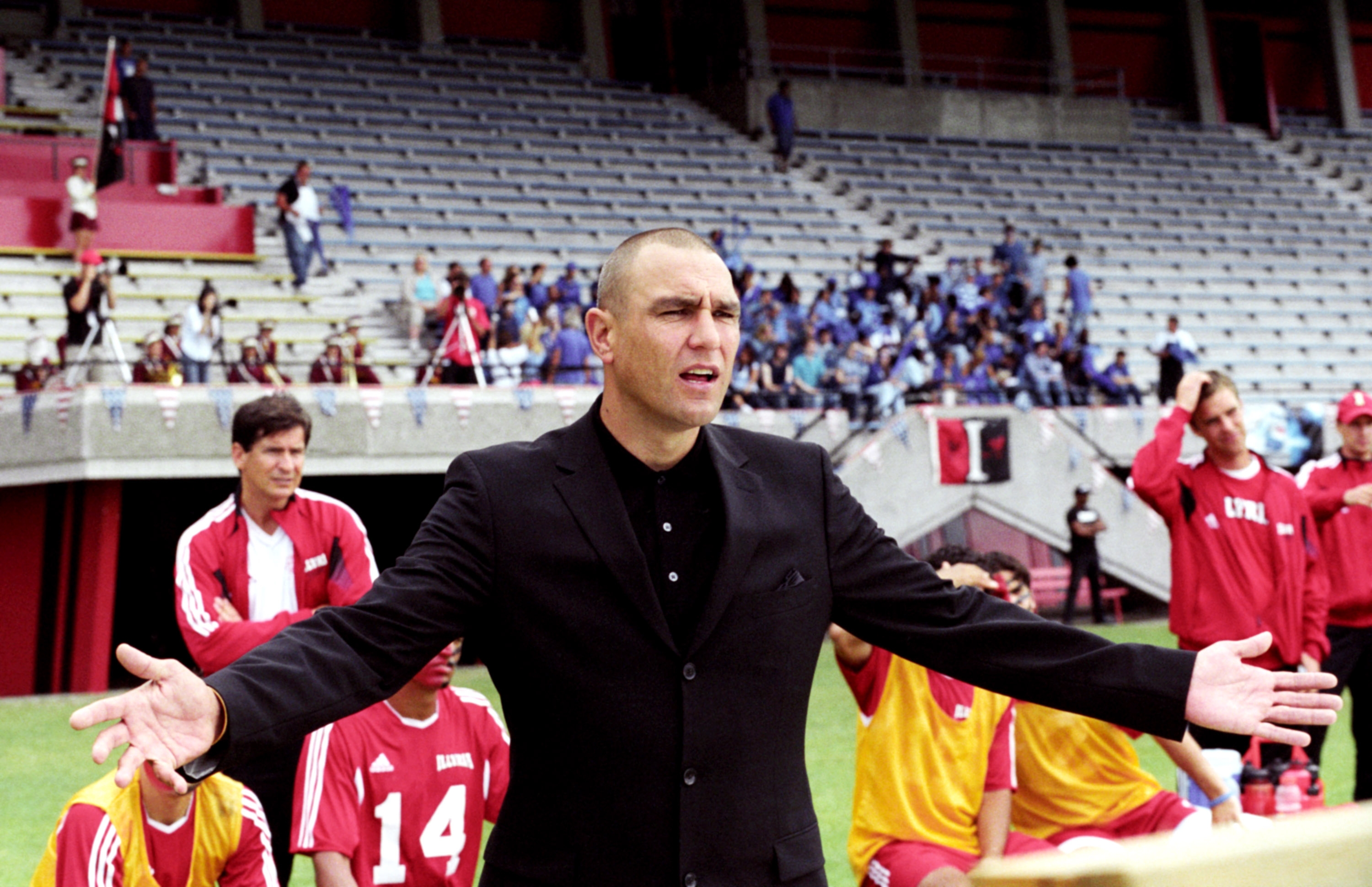 A person in a suit gestures with arms open on a soccer field, surrounded by players in jerseys, with spectators in a stadium background