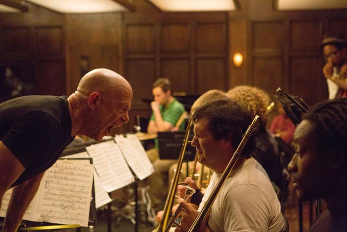 A man intensely directs a seated band in a rehearsal room, focusing on a trombone player