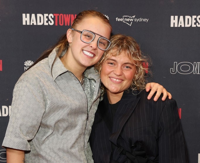 Two people smiling and posing on the "Hadestown" red carpet. One wears a patterned suit; the other a pinstriped outfit