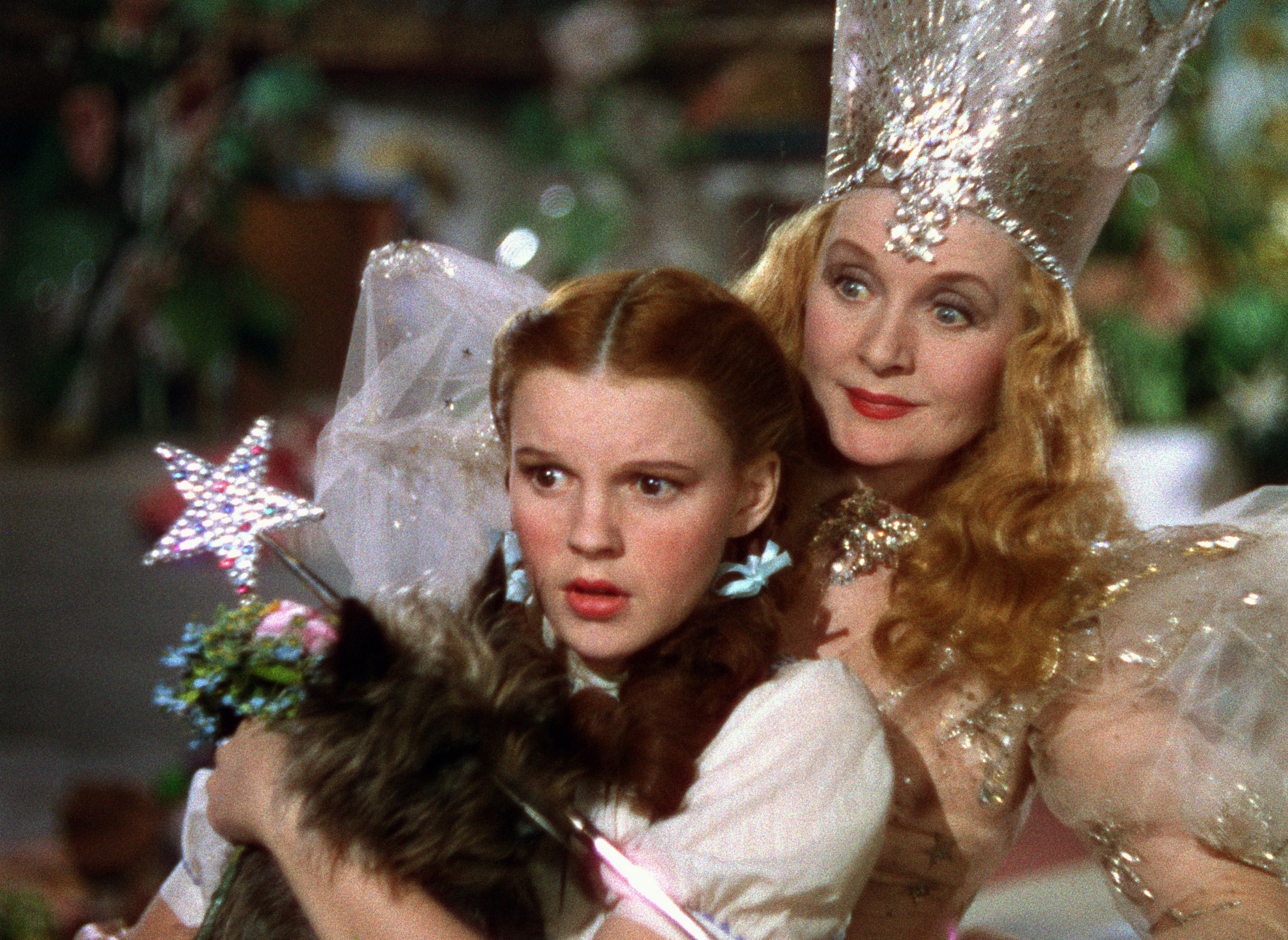 A young girl, holding a dog, looks surprised next to a woman in a sparkling dress and tall jeweled crown, holding a wand