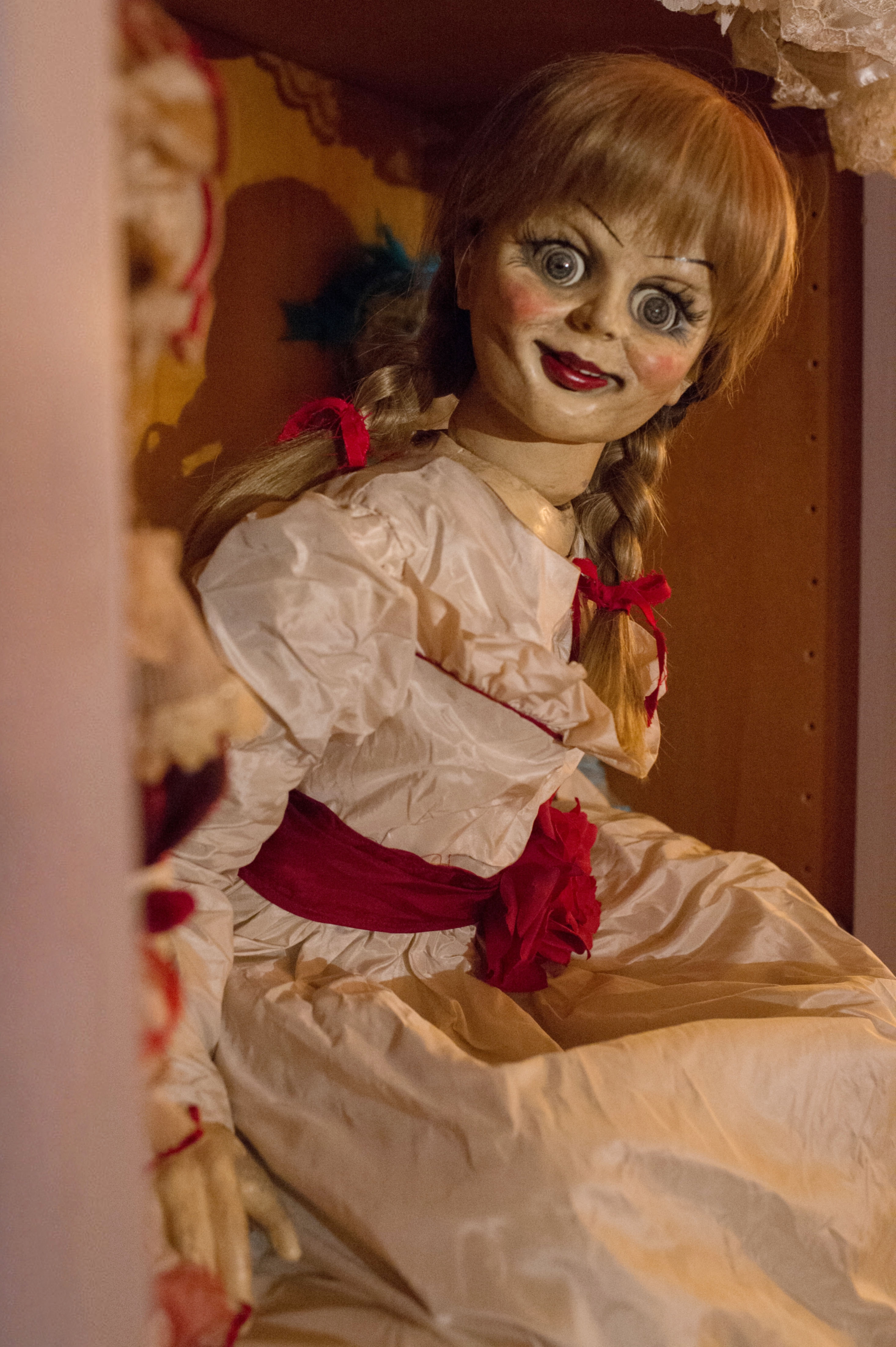 A vintage doll with pigtails and a wide-eyed expression, wearing a frilled dress with a red sash, sits on a wooden shelf