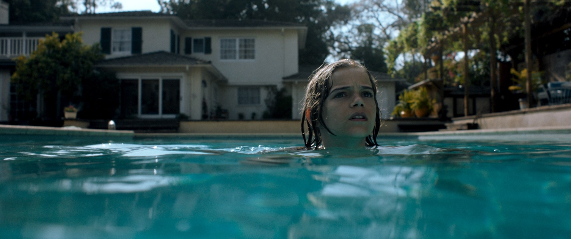 Person swimming in a pool, with a focused expression, against a backdrop of a two-story suburban house and trees