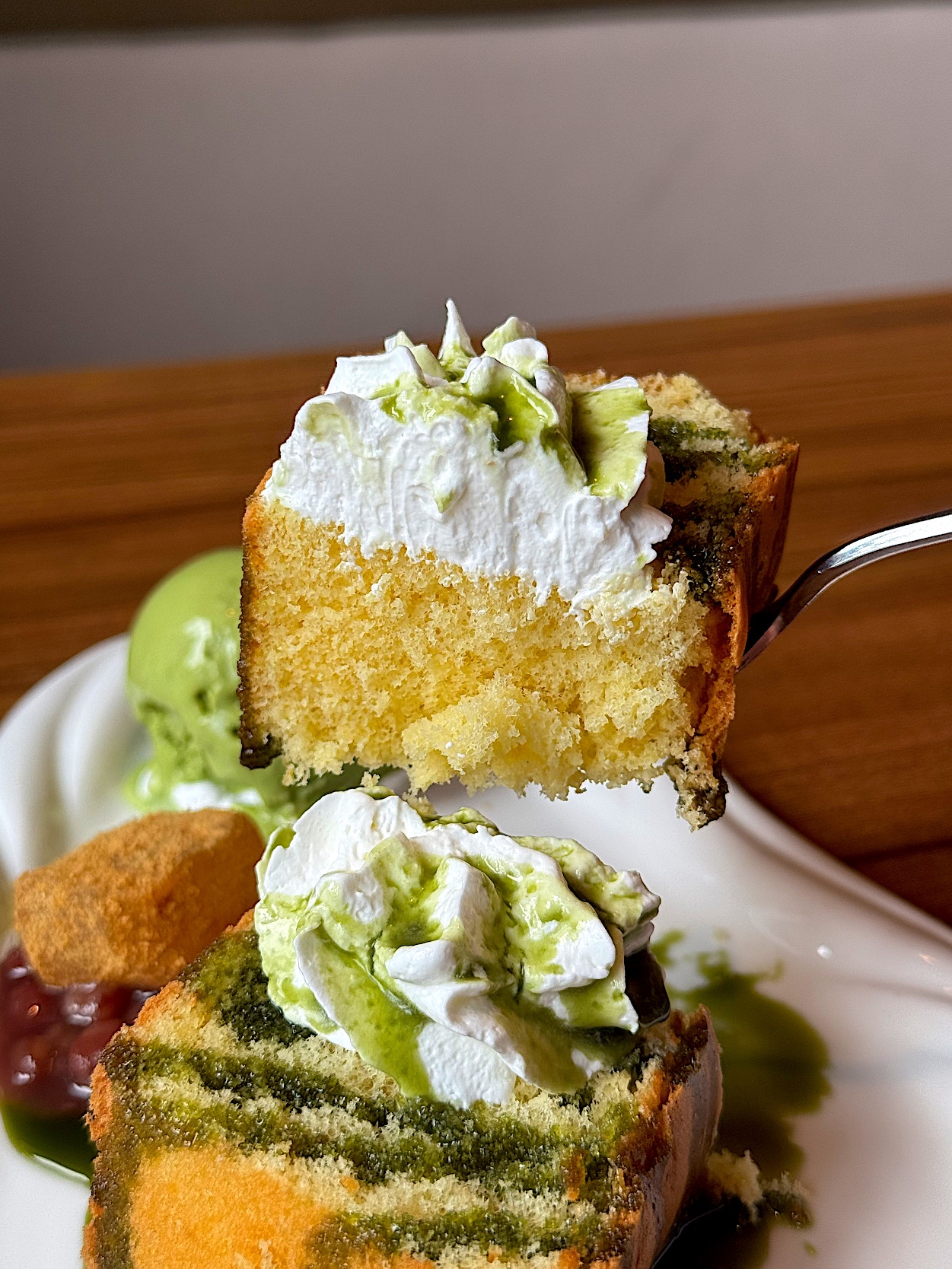 Slice of layered cake with cream and green drizzle, held on a fork. Accompanied by ice cream and a small dessert on a white plate