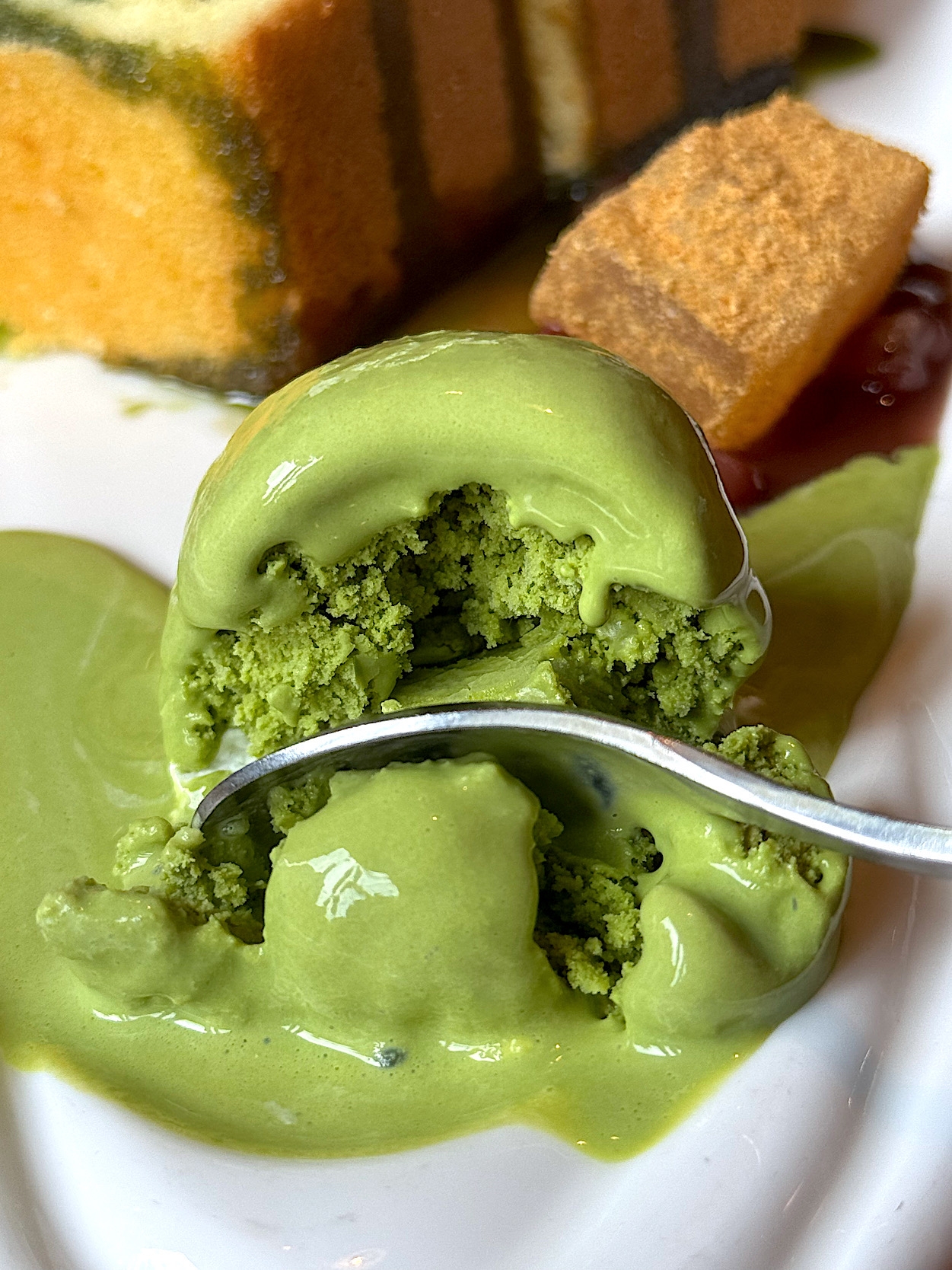 A spoon scoops matcha ice cream on a plate with a slice of layered cake and a cube of soft dessert nearby