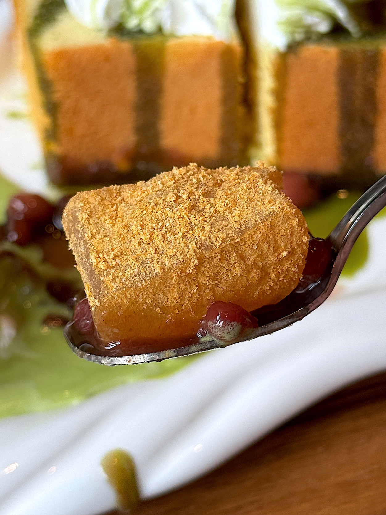 A close-up of a spoon holding a soft dessert covered in powdered topping, with a cake blurred in the background