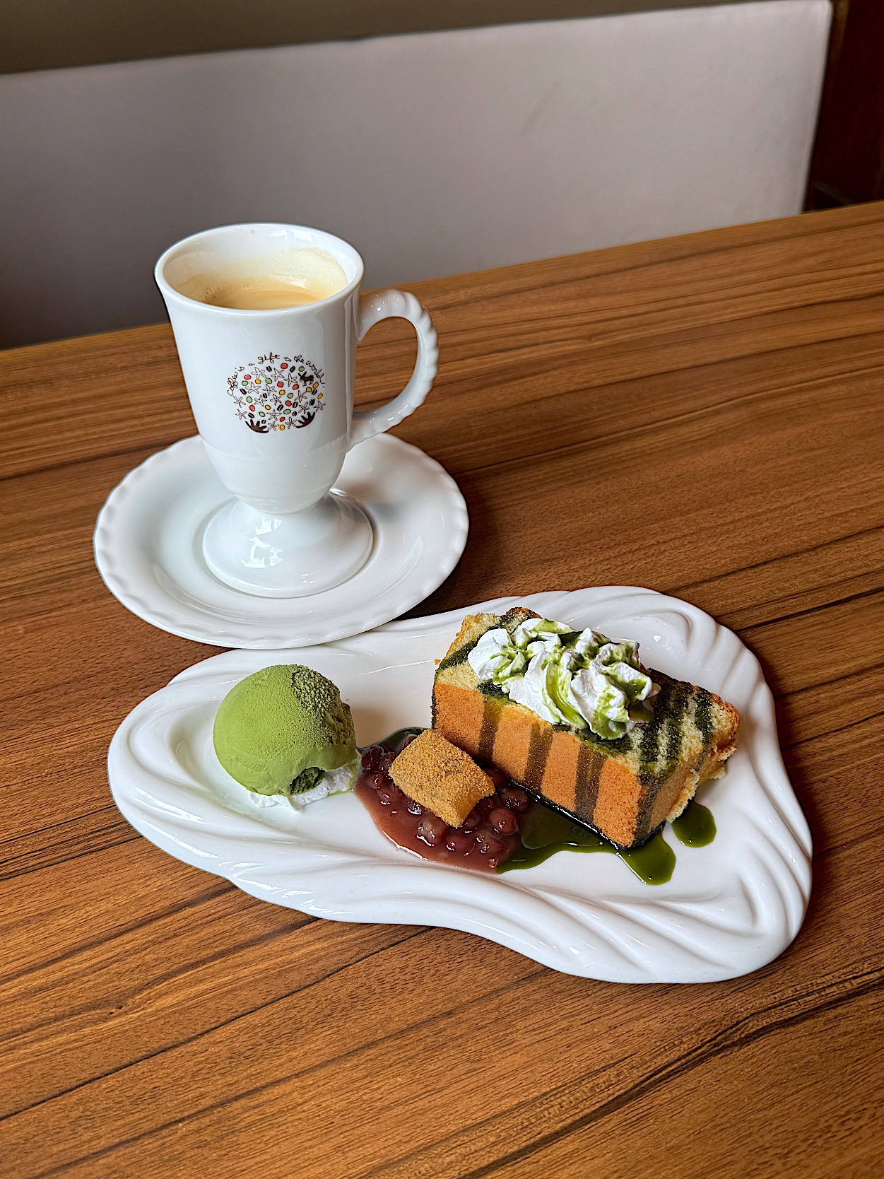 A dessert of green tea cake topped with whipped cream, matcha ice cream, and red bean, served with a cup of coffee on a wooden table