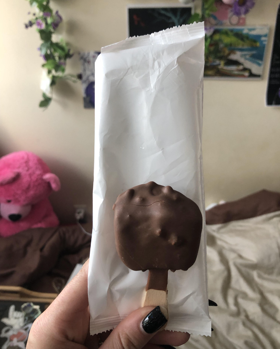Ice cream bar coated in chocolate held by a person with black nail polish. Background includes a plush bear and wall decorations