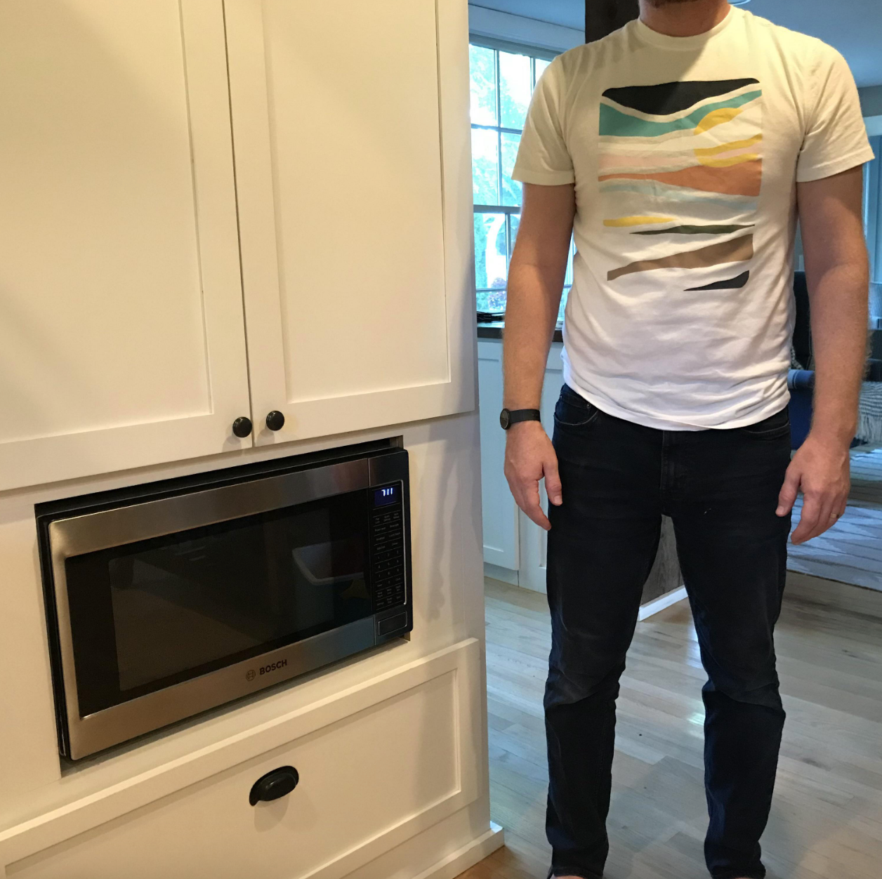 Person wearing a graphic t-shirt and jeans stands beside a microwave built into a white cabinet