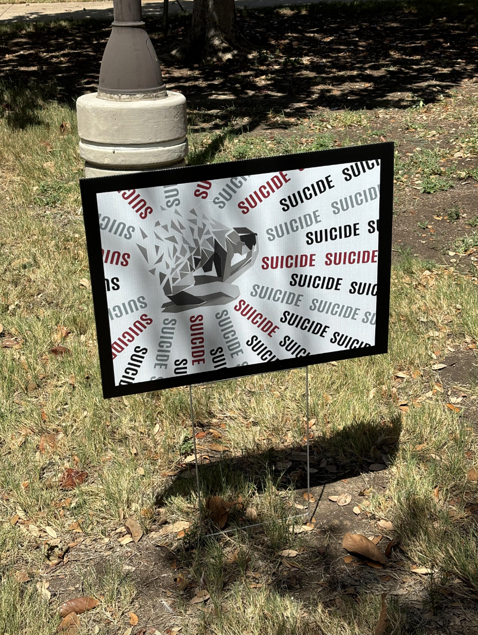 Sign on grass shows fragmented head with repeated words "Suicide" in red and black, highlighting mental health awareness