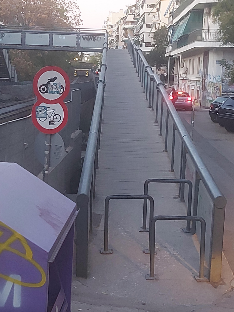 Pedestrian bridge with steep incline and narrow entrance blocked by bike restriction signs, over a city street with cars