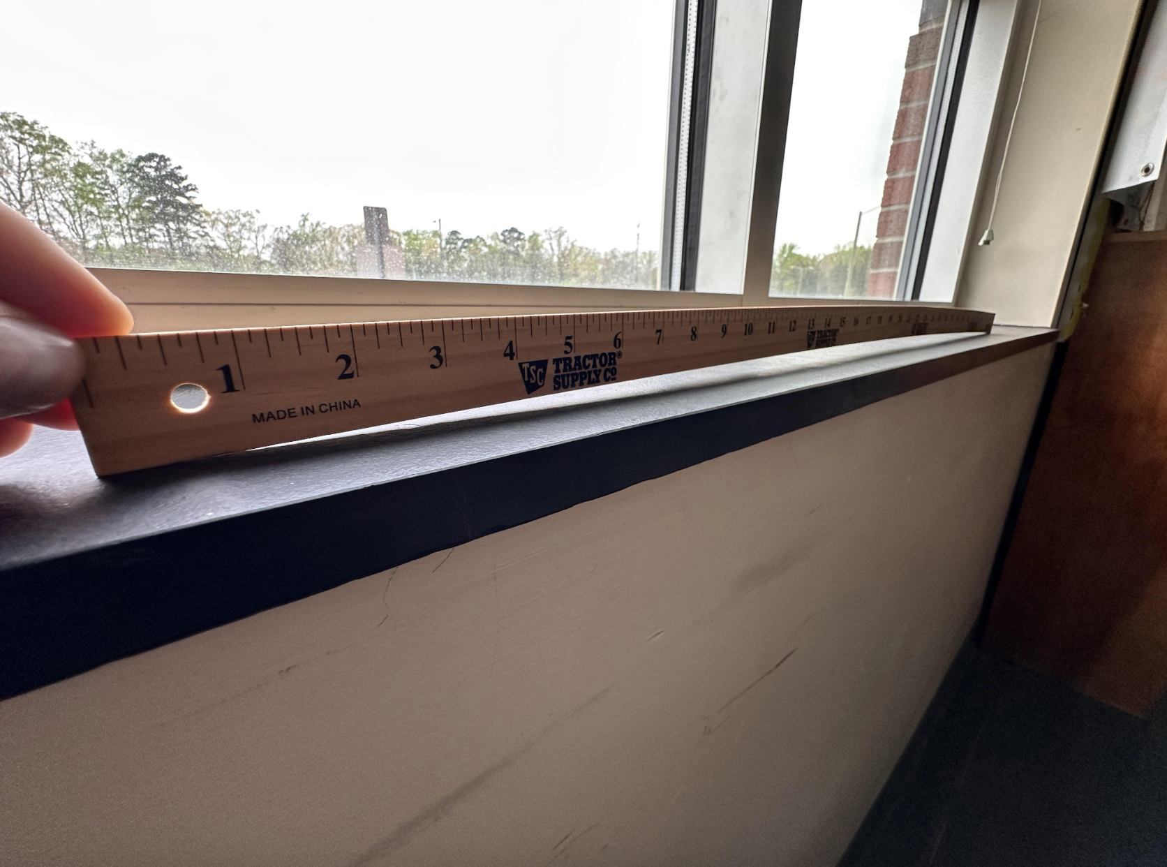 A wooden ruler is held horizontally against a window ledge to measure its width. Trees and a cloudy sky are visible outside the window