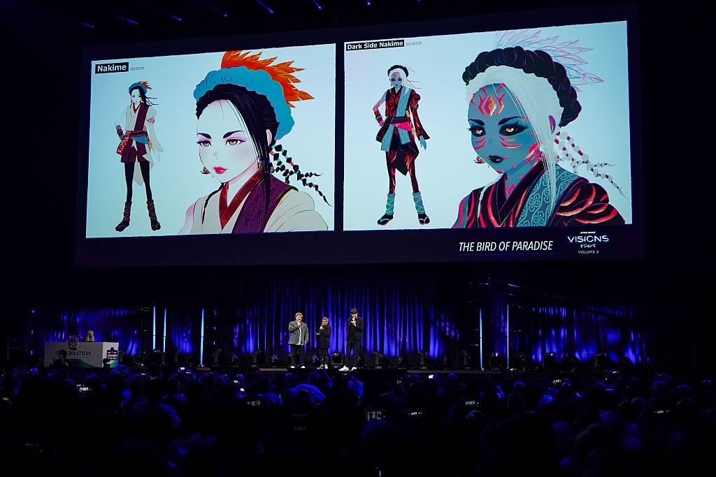A large audience watches a presentation of vibrant animated characters on a screen, featuring "Naksim" and "Dark Side Naksim" from "The Bird of Paradise."