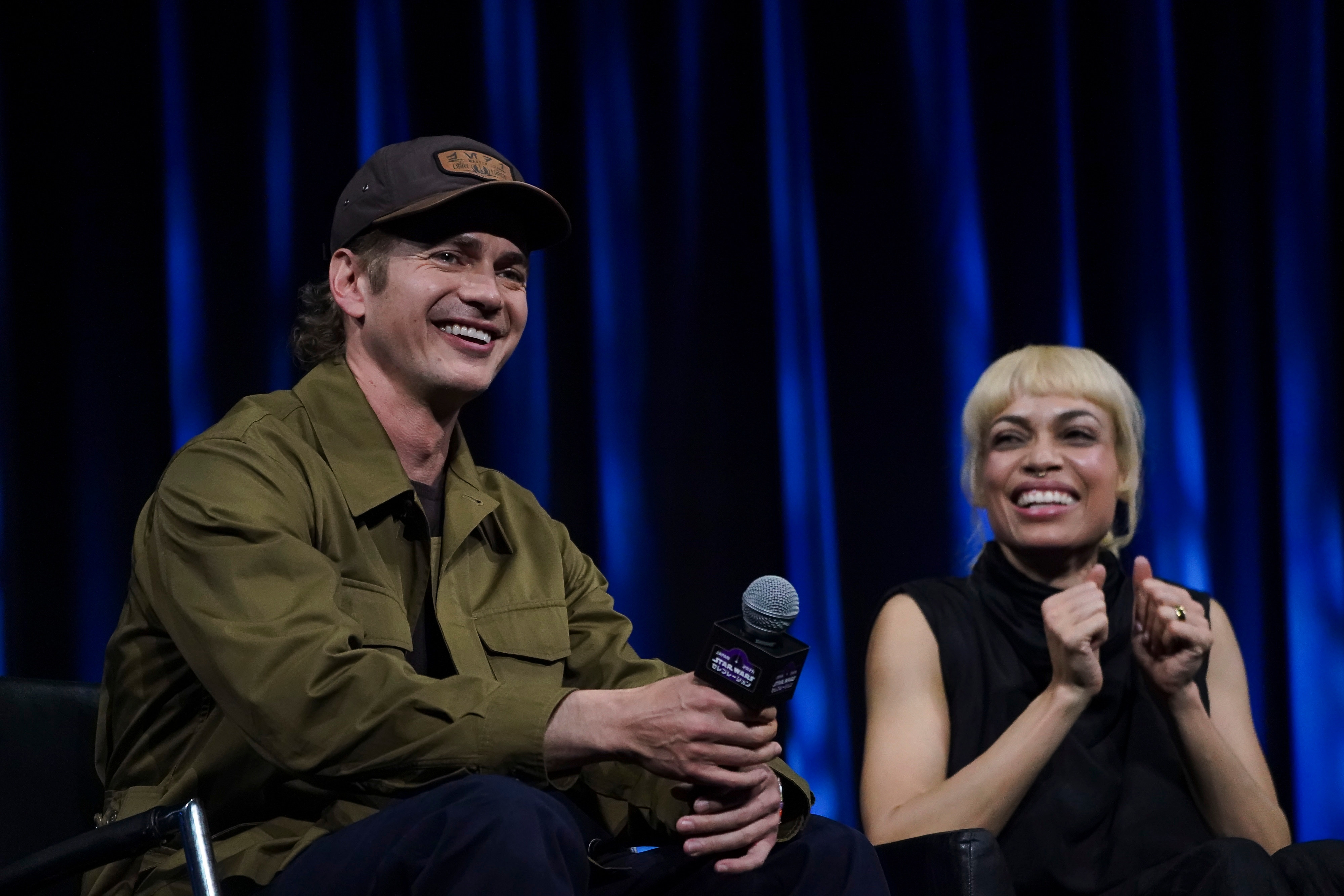Two people sitting and smiling on stage during a panel. One holds a microphone, wearing a casual jacket and cap; the other is in a sleeveless top