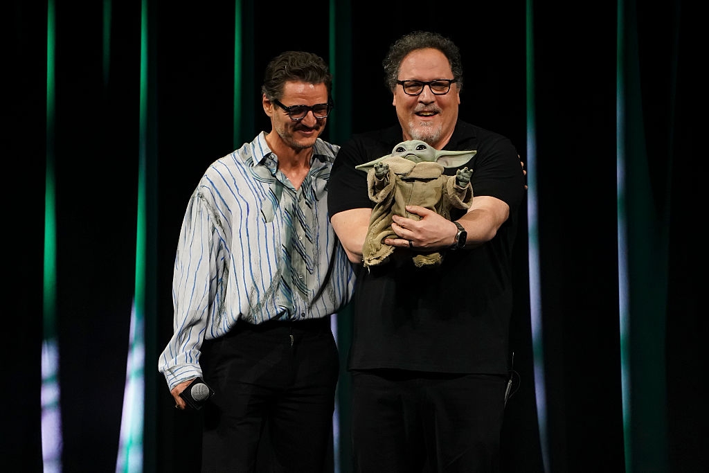 Two men on stage, one holding a puppet resembling Baby Yoda, smiling at an event. The other wears a striped shirt and glasses