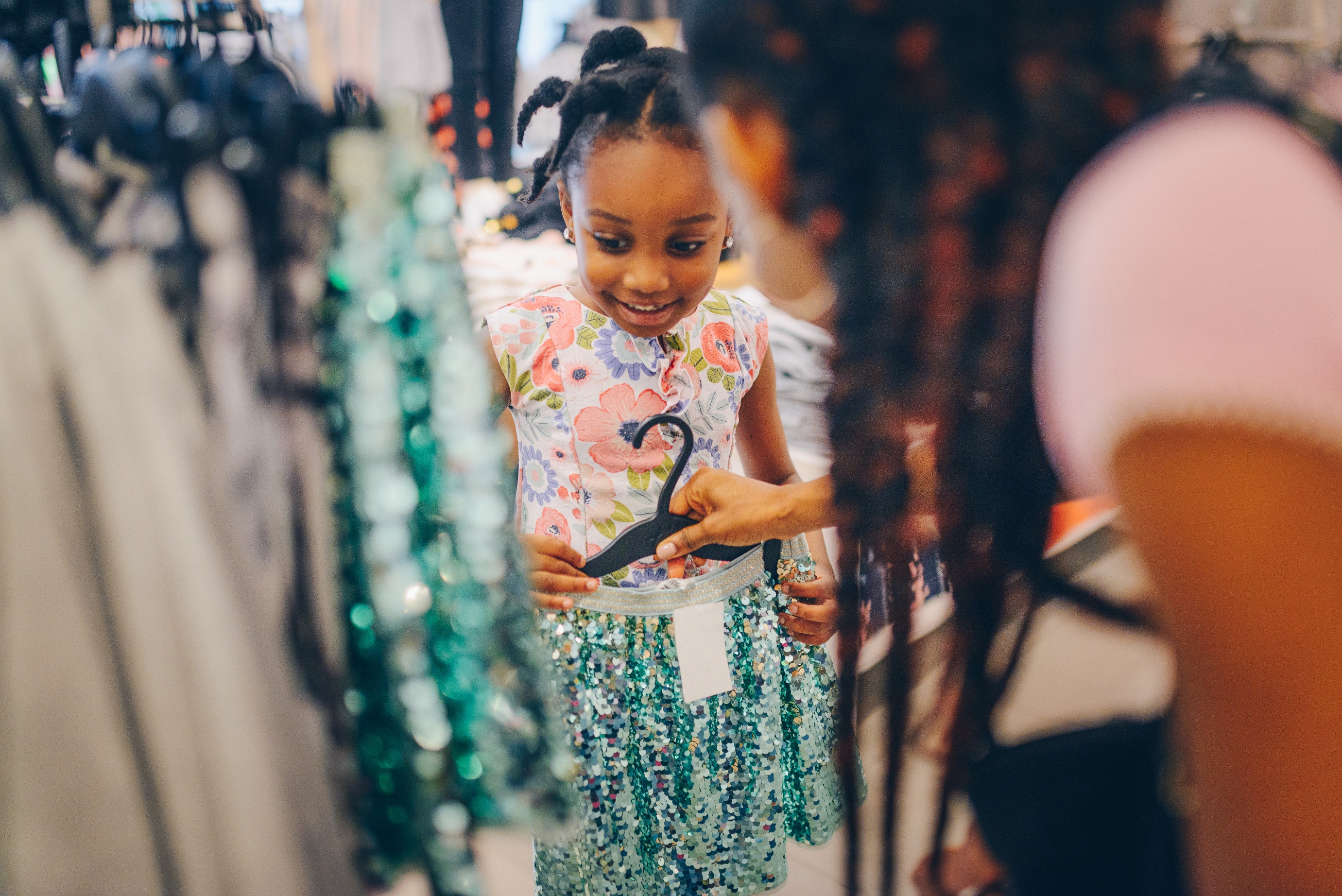A young girl smiles while trying on a sparkly skirt, assisted by an adult. They are in a clothing store, surrounded by more clothes