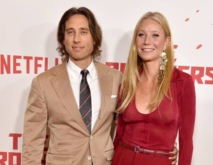 Two individuals pose on a red carpet. One wears a beige suit with a striped tie, the other in a fitted red dress. They smile at the camera