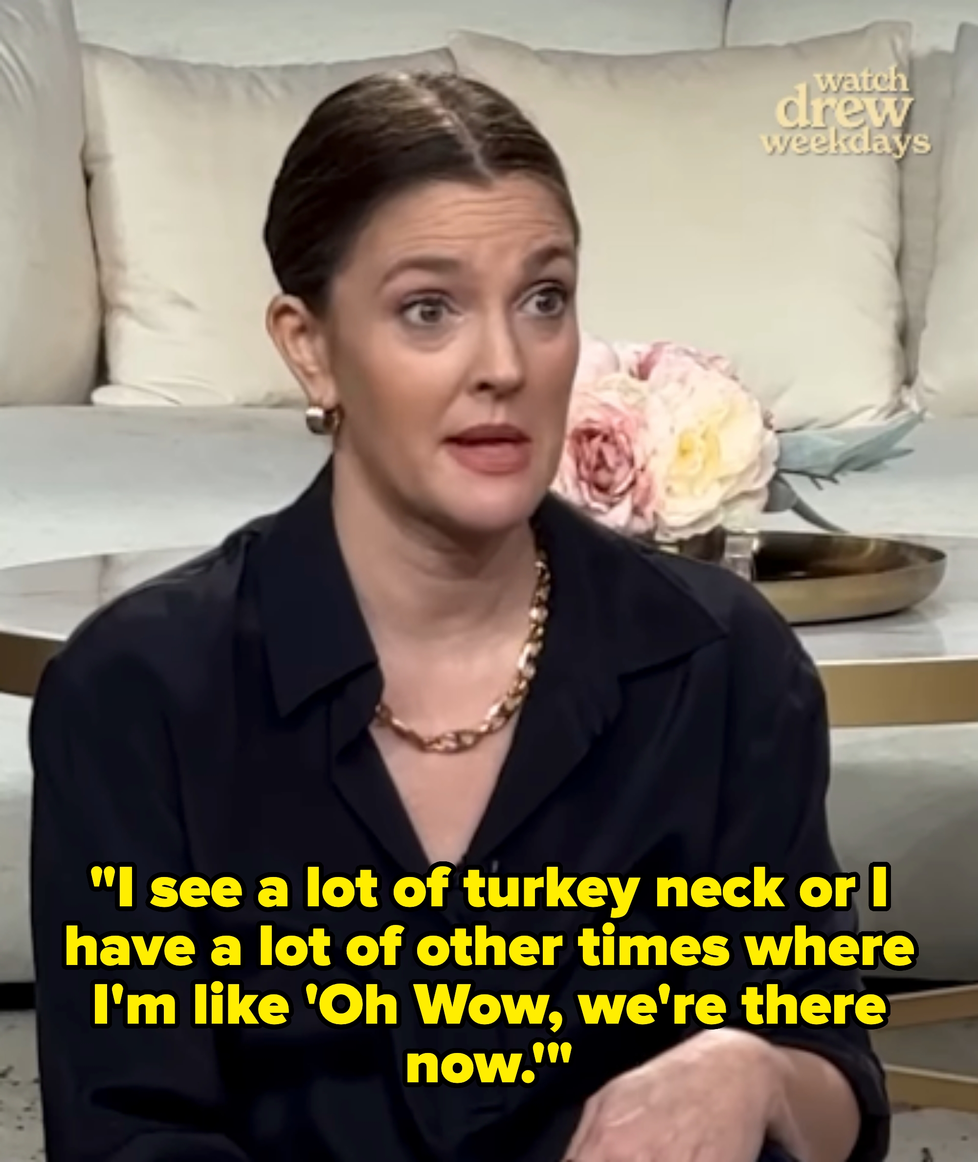 Person with slicked-back hair in a dark blouse, sitting on a couch, speaking