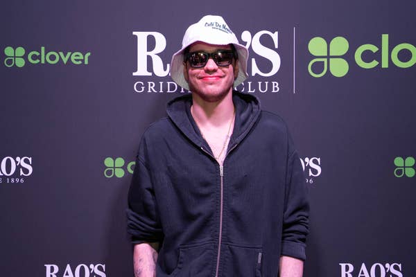 Pete Davidson in a hoodie, sunglasses, and a cap poses at an event backdrop with logos
