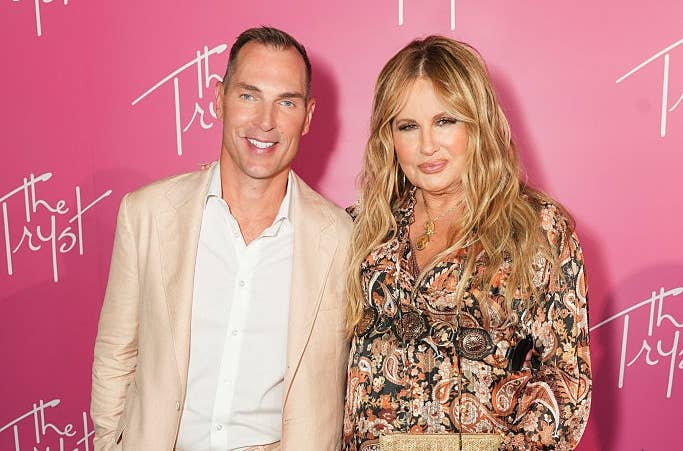 A man in a light suit and a woman in a patterned dress stand together on a patterned floor, posing for a photo in front of a pink backdrop with "The Trust."