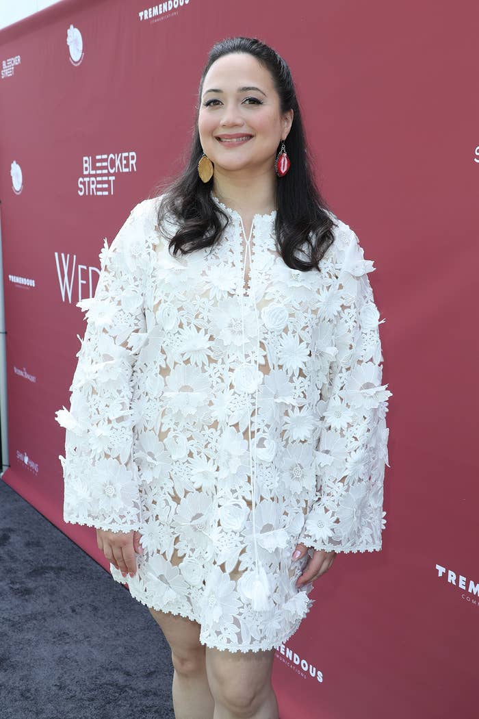 Lily Gladstone on a red carpet wearing a floral textured dress with long sleeves, smiling at the camera