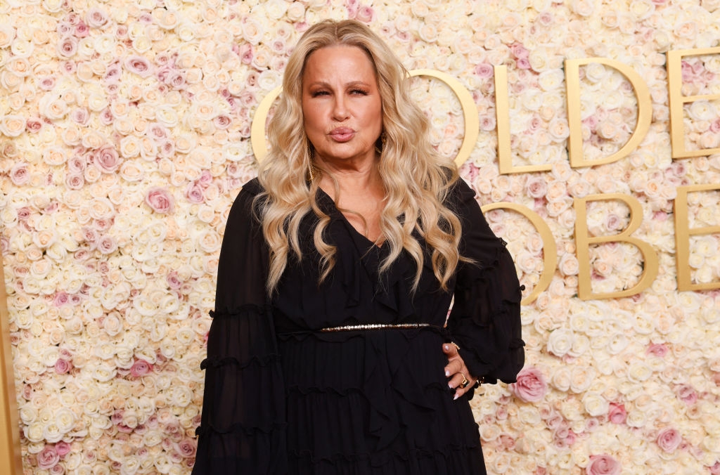 Person poses in front of a flower wall, wearing a black, ruffled dress with long blonde hair styled in waves